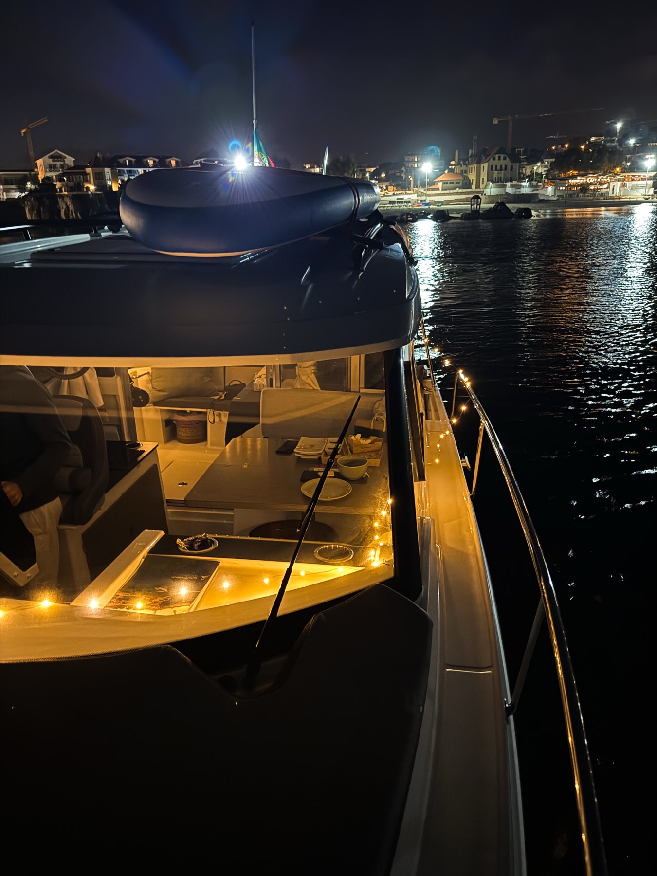 Private boat glowing with warm string lights at night in the bay of Cascais, with the town lights in the background